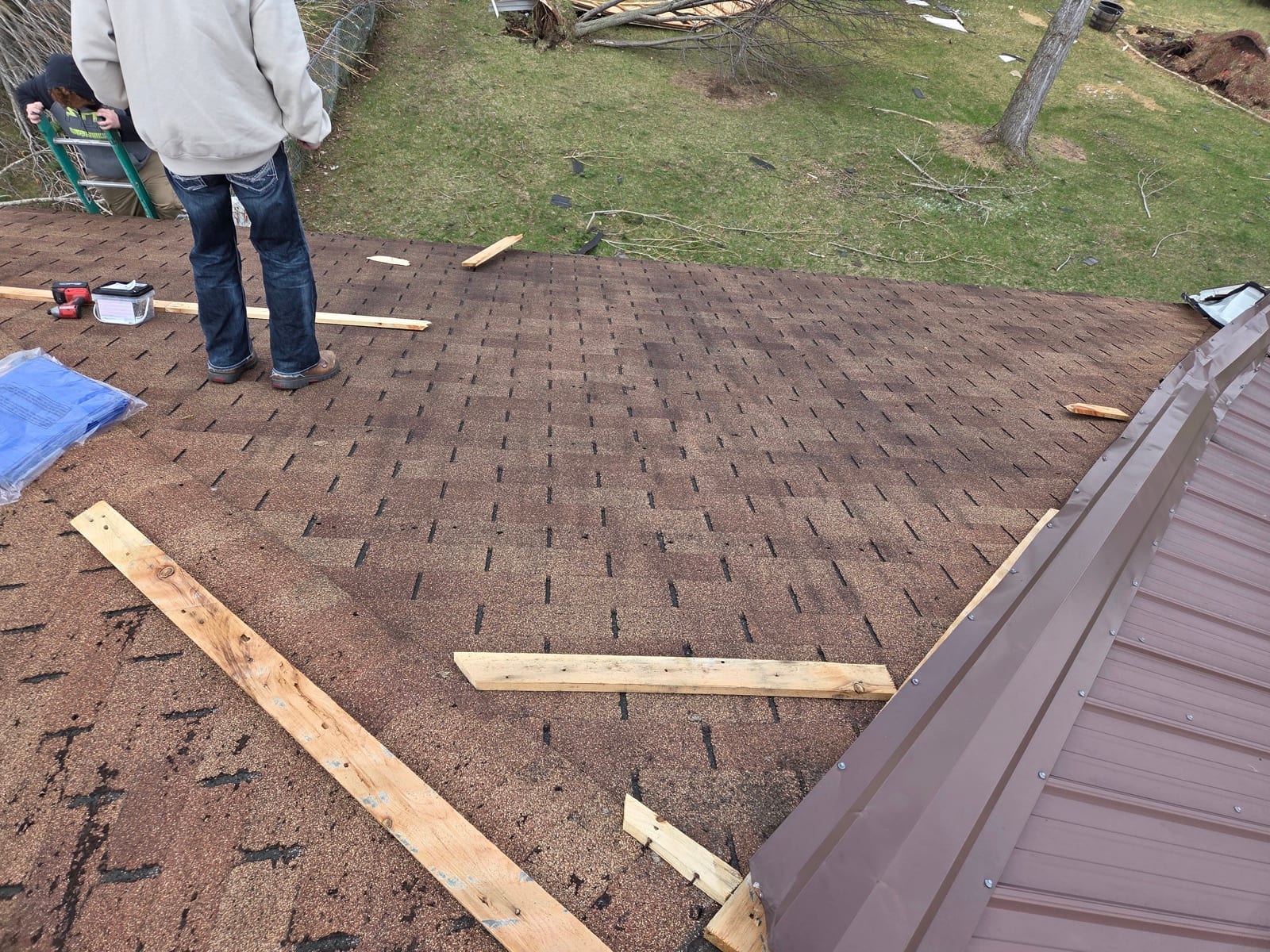 Shield crew members on a roof mid-repair, with storm debris scattered across the yard below.